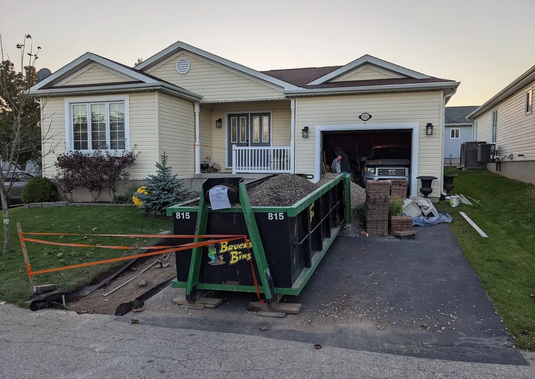 A nearly full disposal bin laying on a driveway, with a sizable trench dug out running alongside the driveway